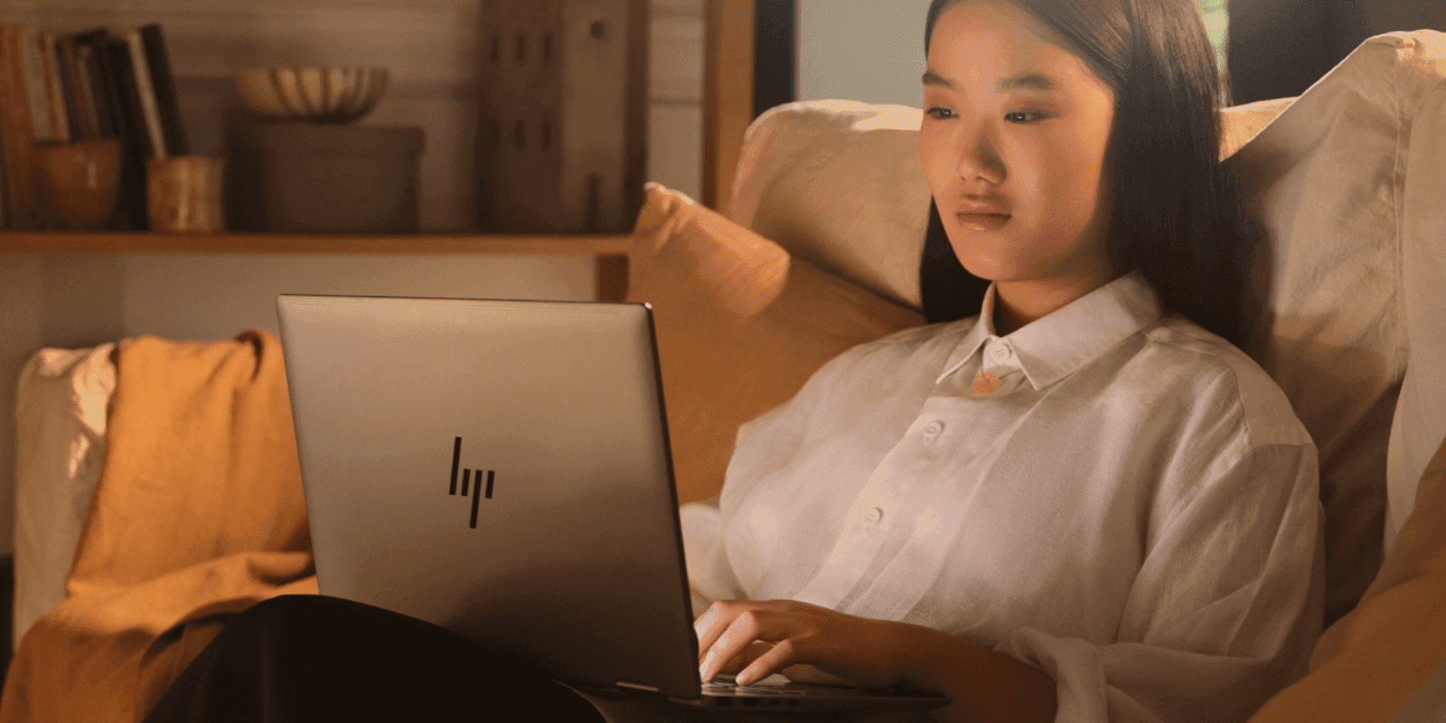 A young Asian woman sits comfortably on a couch, working on an HP laptop in a warmly lit living room with bookshelves visible in the background.