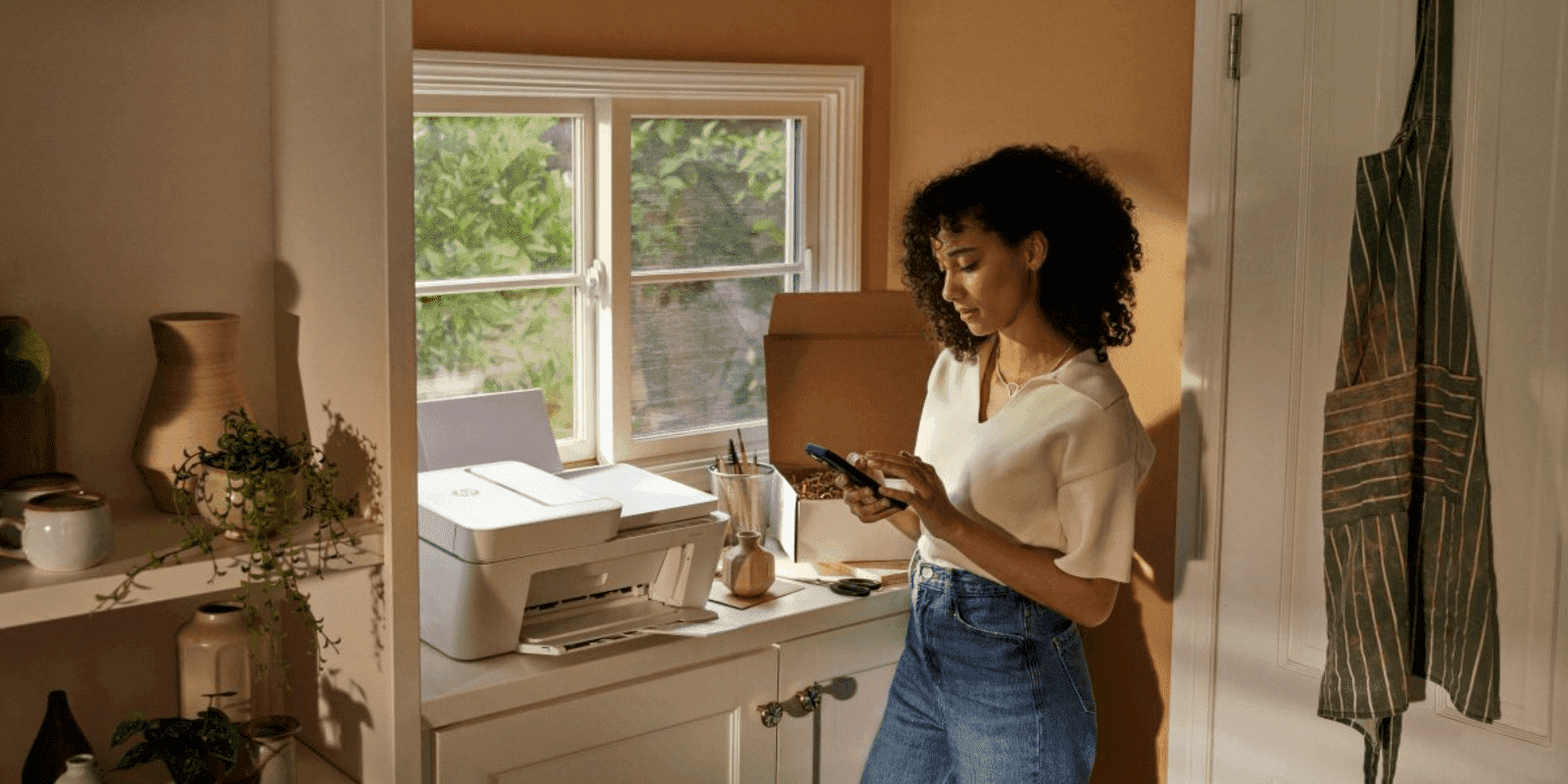 A young woman with curly hair stands in a sunlit home office, using her smartphone near a desk with a printer, surrounded by plants and warm, natural decor.