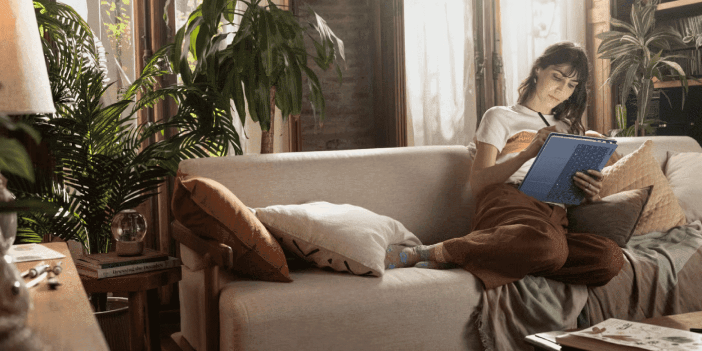 A woman is looking at her HP OmniBook X Flip 14 while relaxing on a beige couch in a cozy, plant-filled living room with natural lighting and bookshelves in the background. Share HP OmniBook X Flip 14: Is It the Best Convertible Laptop for Modern Professi